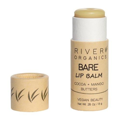 A beige cylindrical tube of River Organics Bare Vegan Lip Balm with the cap off, revealing the balm stick. The tube is labeled with "Cocoa + Mango Butters" and "Vegan Beauty," emphasizing its appeal as an ultra-hydrating, vegan lip balm. A subtle leaf design adorns the bottom. Net weight is 0.28 oz (8 g).